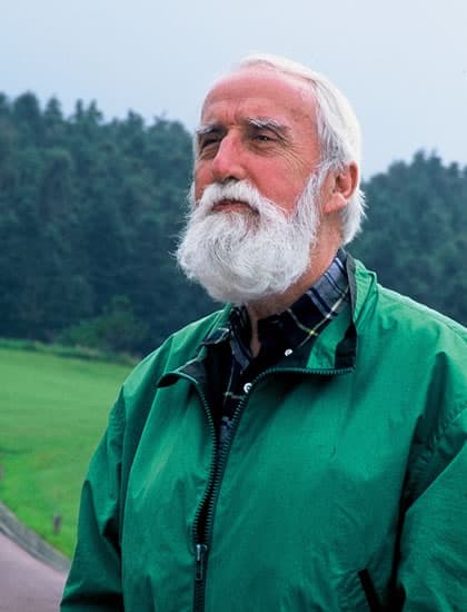 Portrait of golf course architect Desmond Muirhead, white-bearded in a green jacket, standing before a forested hillside