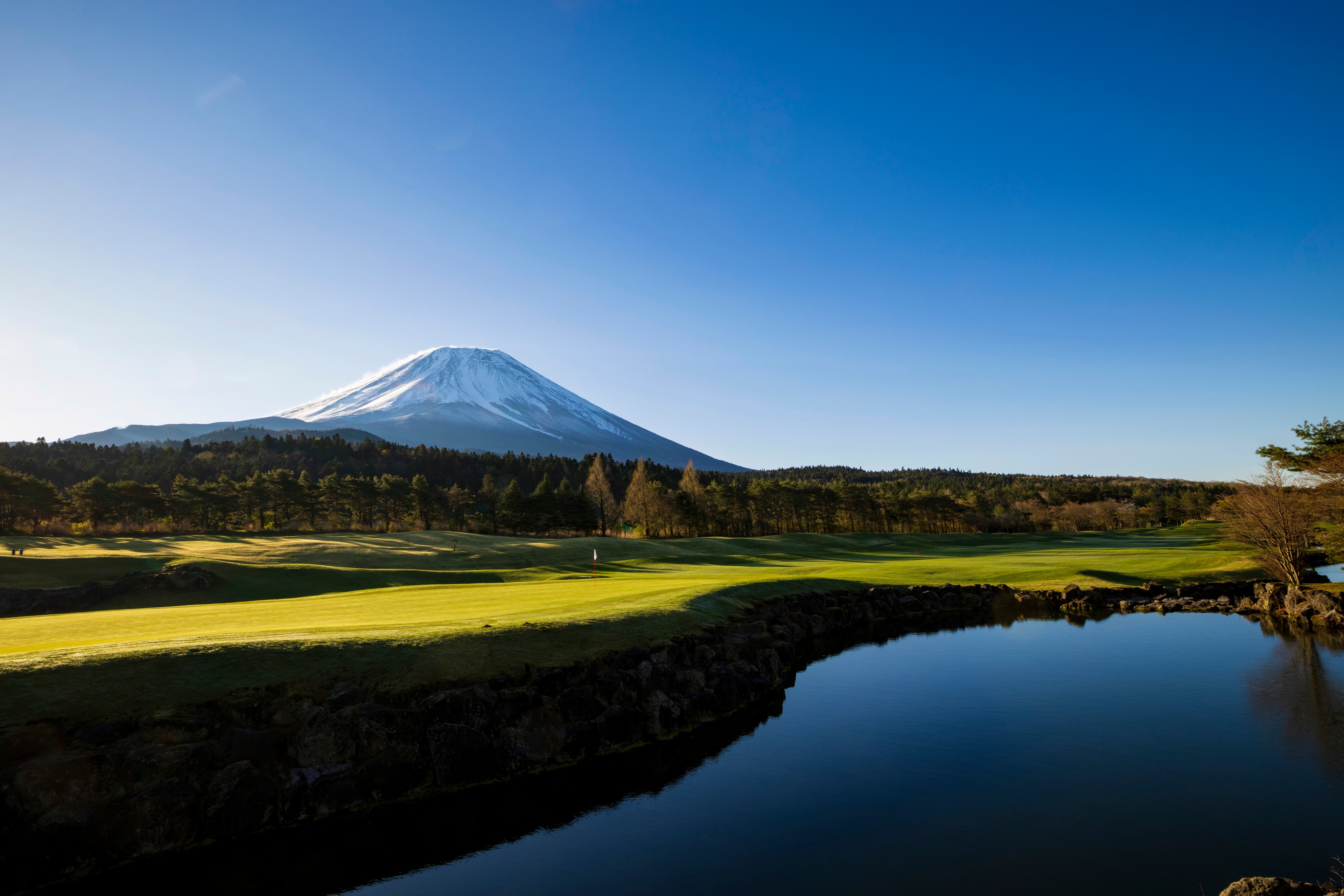 Snow-capped Mt. Fuji rising above a sunlit green and a still pond at Fuji Classic
